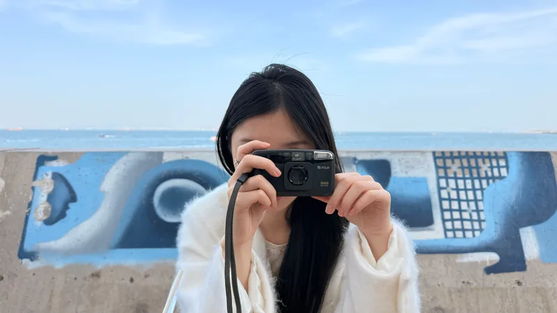 她在海边举着相机对向镜头，身后是蓝色墙绘