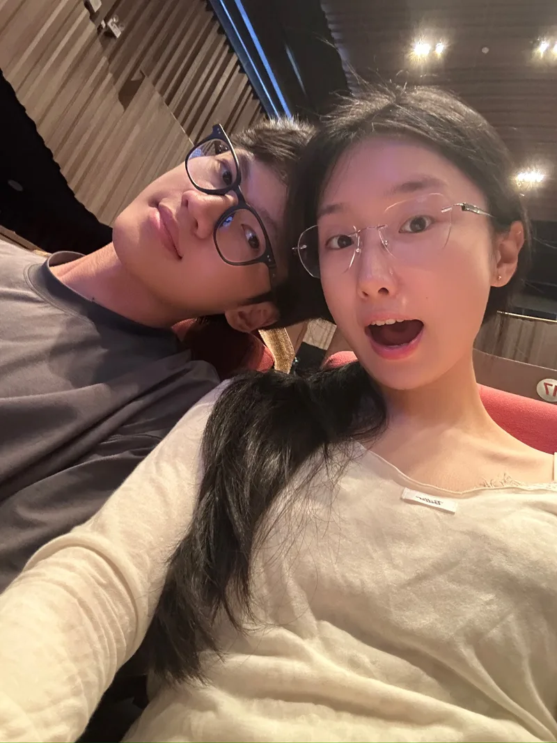 音乐会观众席上，两人戴着眼镜挨在一起自拍，身后是红色座椅和木色墙面