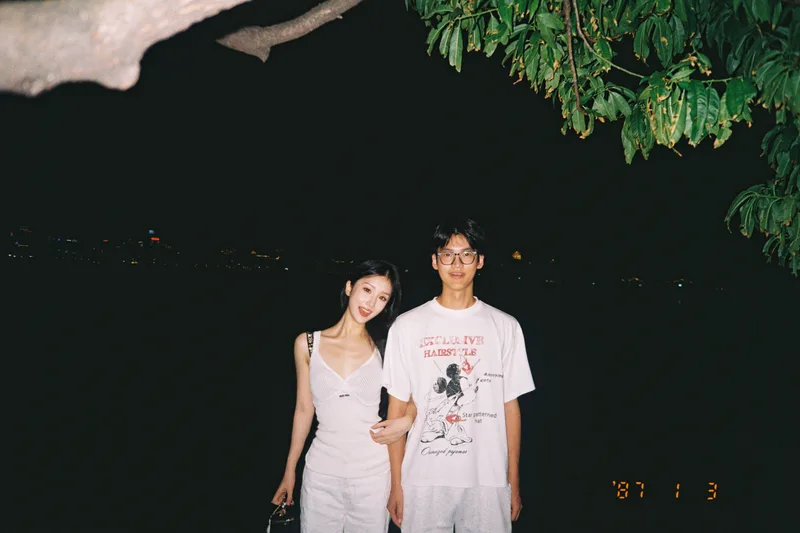 夜晚的西湖边，两人穿着浅色衣服站在树下，她挽着他的手臂，身后是湖面与远处灯光