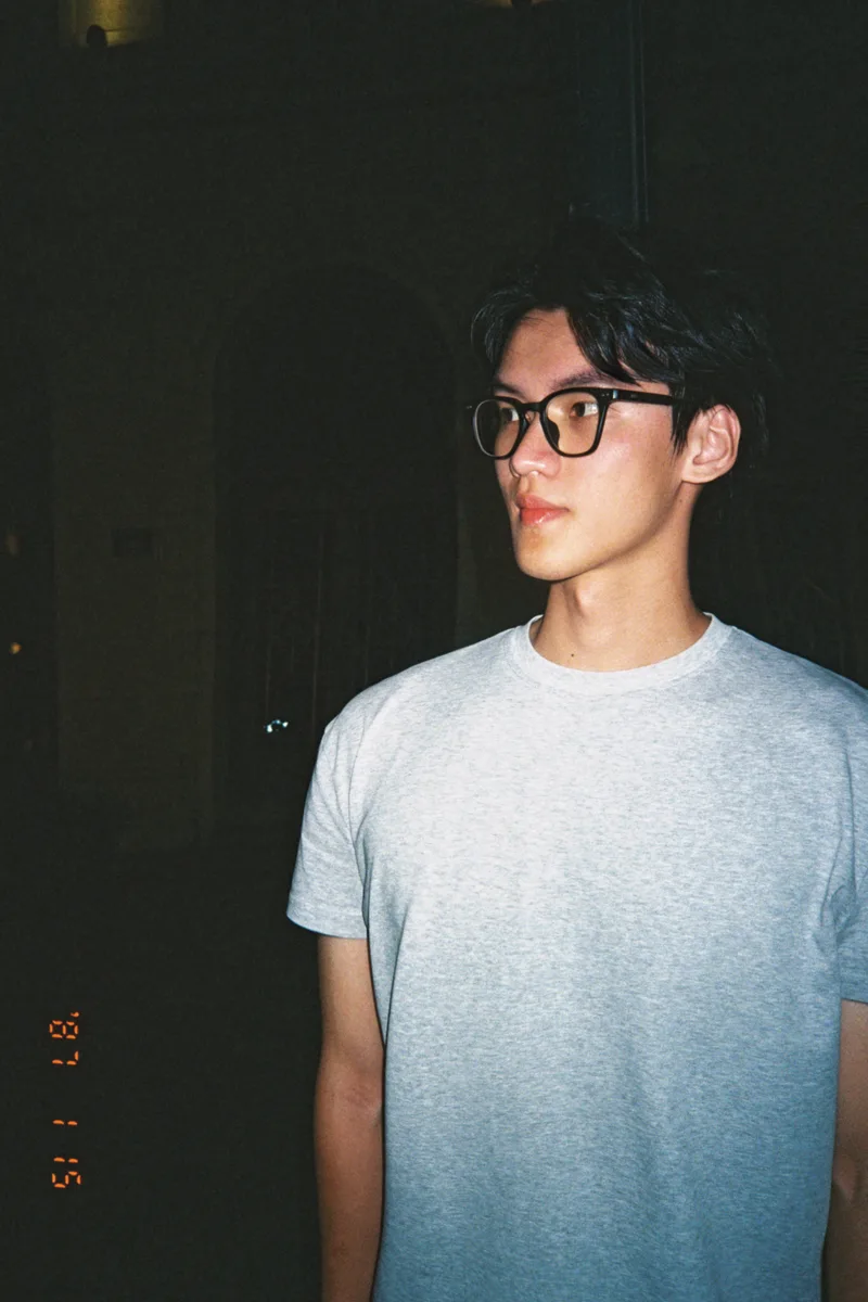 夜晚街边，他戴着黑框眼镜，穿着灰色上衣望向一旁