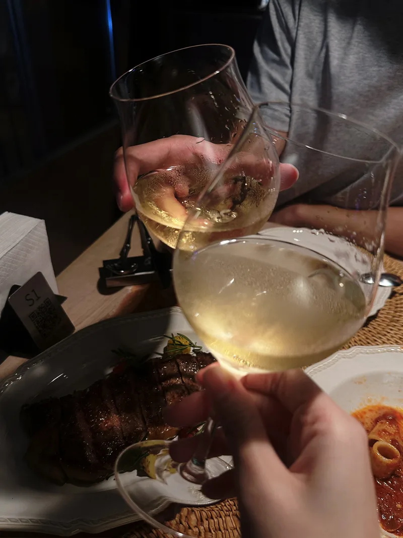 两只盛着浅色酒液的高脚杯在餐桌前碰在一起，桌上摆着晚餐