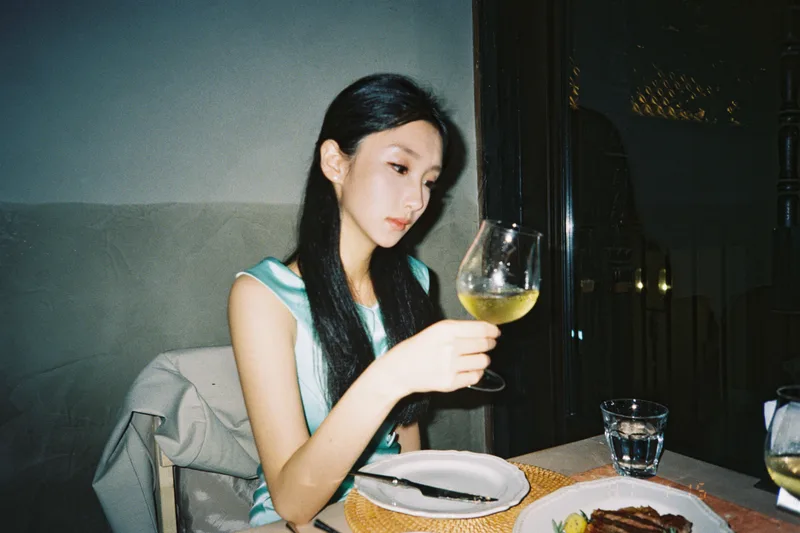 她穿着浅绿色裙子坐在餐馆窗边，手里举着高脚杯