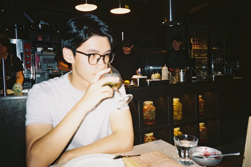 他穿着灰色上衣在餐馆里端起高脚杯，身后是开放式厨房