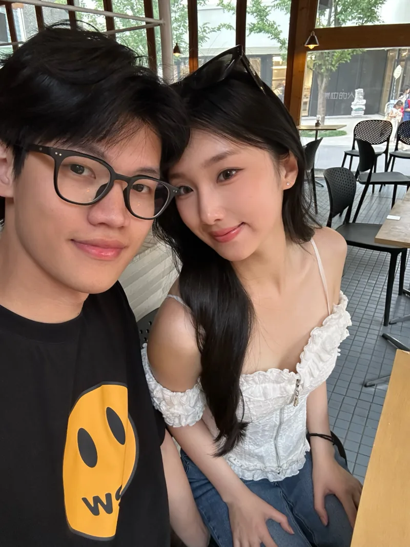 两人坐在咖啡店木窗旁,头靠在一起自拍,窗外有树