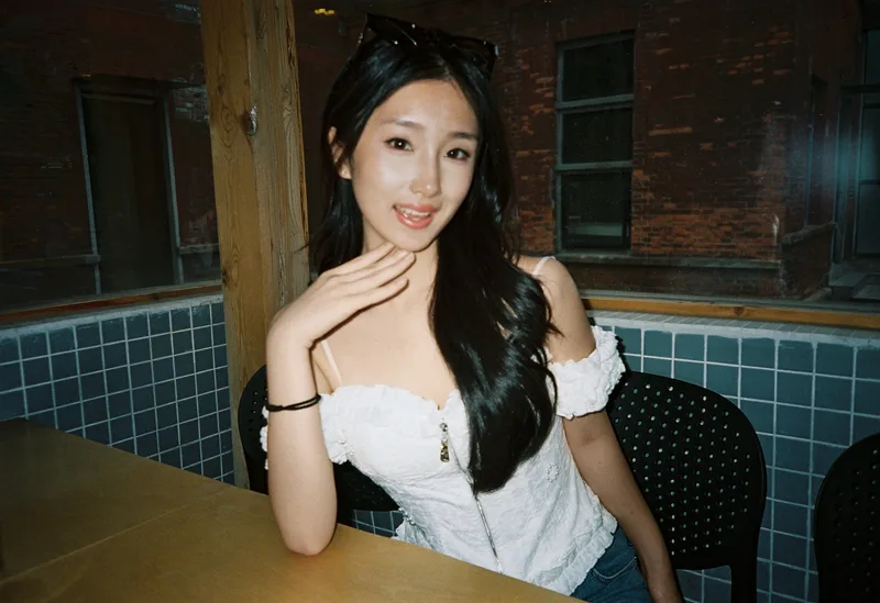 她穿白色上衣坐在咖啡店木窗与蓝色方砖旁，抬手轻托下巴