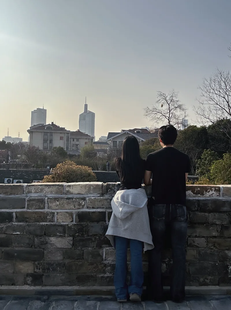 两人并肩站在砖墙旁,背对镜头望向南京的街景