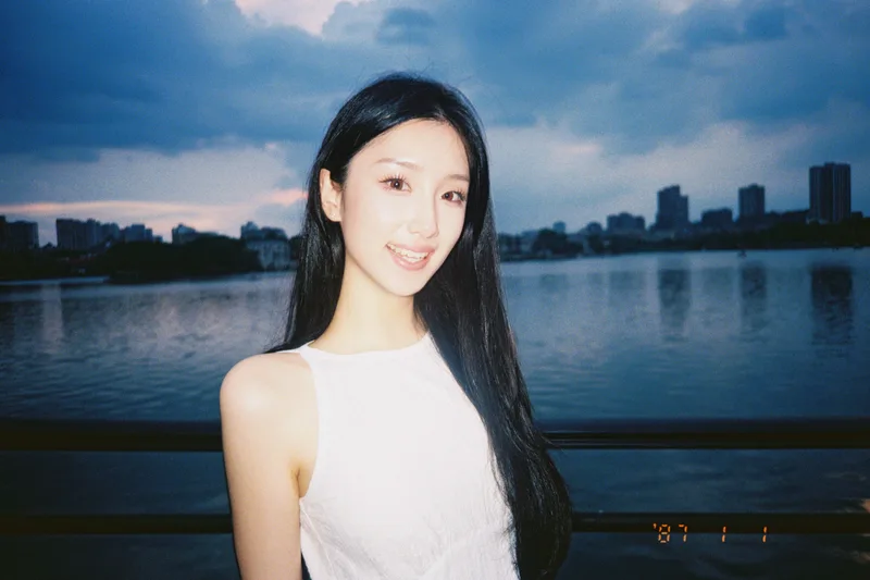 她站在百家湖旁微笑，背后是湖面和蓝灰色的天空