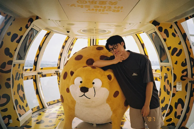 他站在同一个黄色大玩偶旁，抬手托着下巴看向镜头