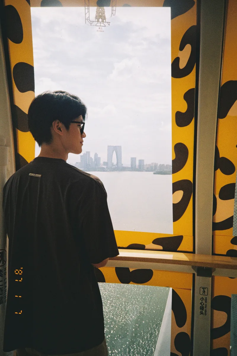 他戴着墨镜站在摩天轮窗边，望向金鸡湖和远处的建筑