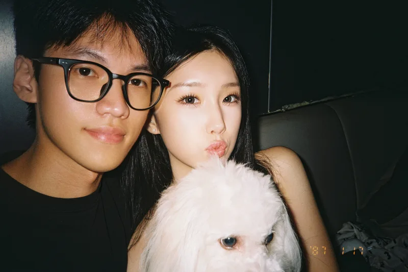 两人在深色卡座前合照，她抱着白色小狗可颂，朝镜头嘟嘴