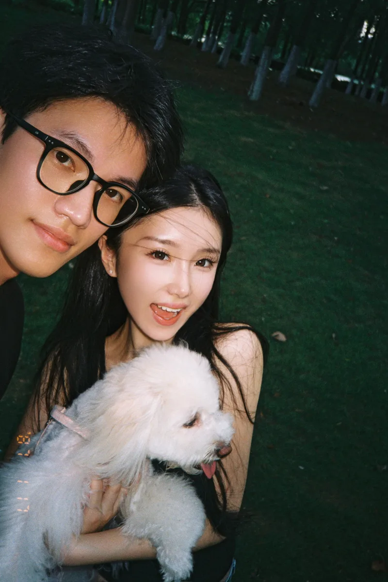两人在户外树木前一起自拍，她怀里抱着白色小狗可颂